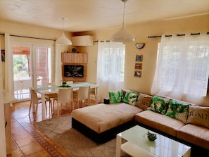 A living room with a sofa, a dining table with chairs, a TV mounted on the wall, and a kitchen area with cabinets.