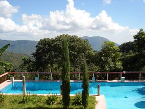 A rectangular swimming pool with a clear blue water surface, surrounded by a red deck and white pillars. In the background, there are lush green trees and a mountain range under a partly cloudy sky.