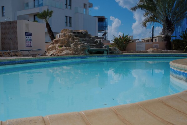 A swimming pool with a rock feature, palm trees, and modern buildings in the background.