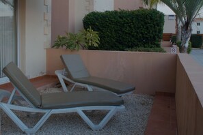 Two lounge chairs with cushions, a small potted plant, and a hedge in a courtyard.