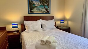 Habitación de hotel con cama, dos mesitas de noche, un cuadro de una playa y dos lámparas.