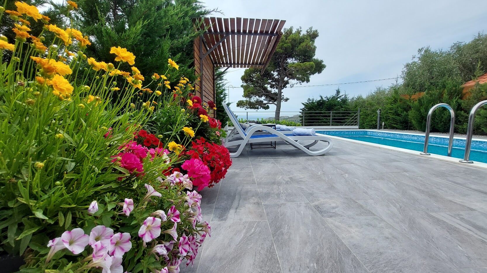 Área ao lado da piscina com pergolado, espreguiçadeiras e diversas flores.