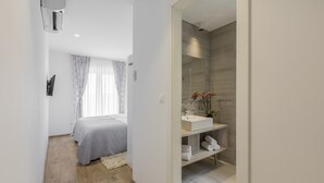 Een moderne slaapkamer met een bed, nachtkastje en een klein tafeltje. Er is een badkamer met een wastafel, spiegel en handdoekhouder.
