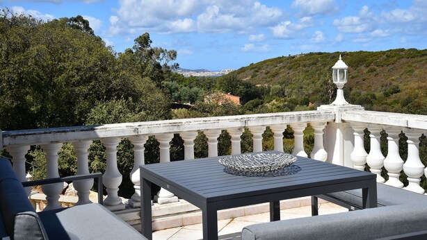 Un balcón con mesa y asientos, con vistas a un paisaje pintoresco con árboles y colinas.