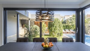 Un espace repas moderne avec une grande table, un panier de fruits et une vue sur une piscine et un coin salon extérieur.