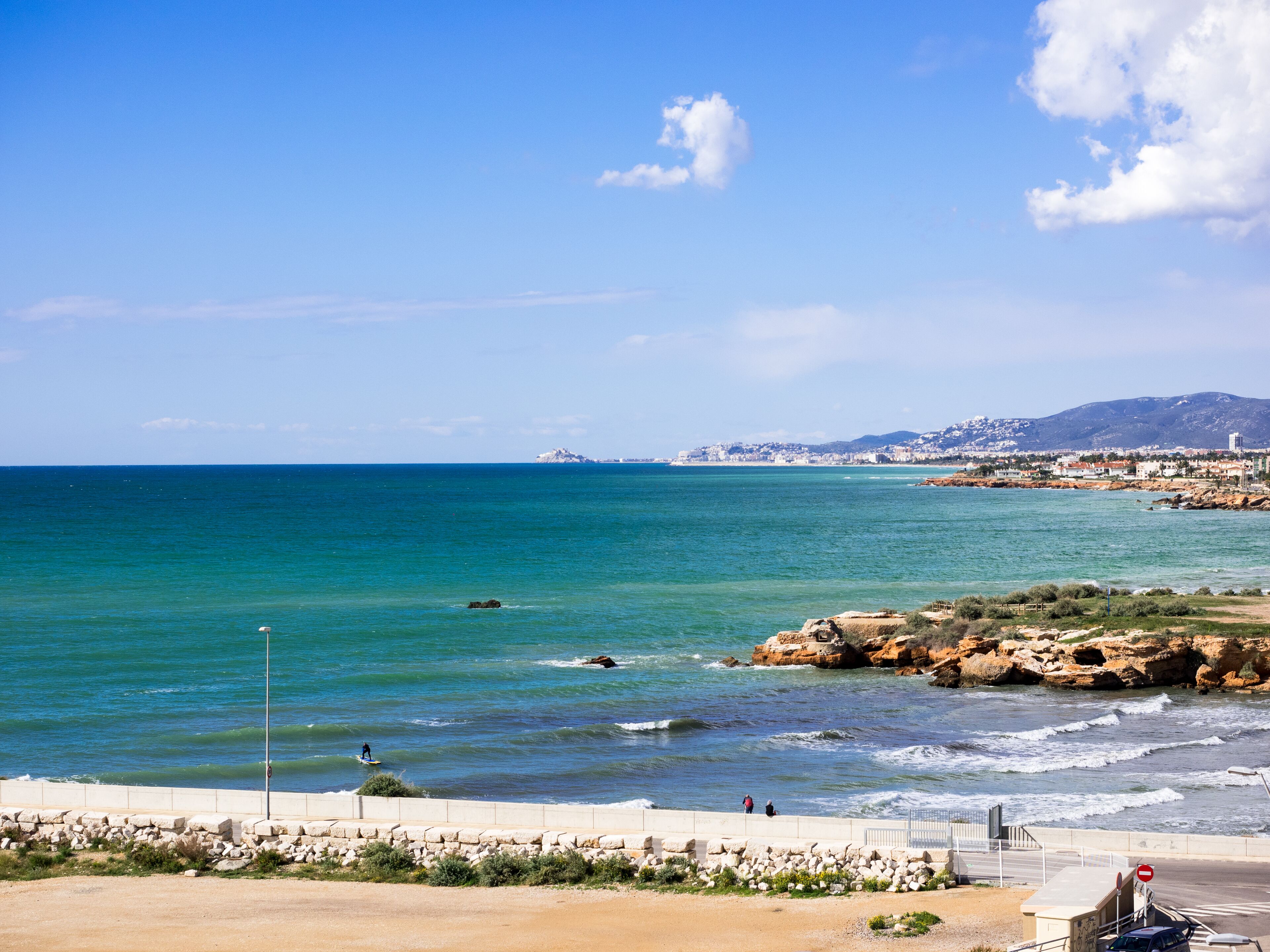 Vista costera con cielo despejado, mar tranquilo, costa rocosa y un paisaje urbano lejano.