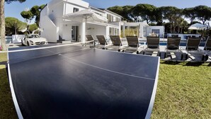 Un court de tennis avec une voiture blanche garée à proximité, un espace piscine avec des chaises longues, et un bâtiment moderne doté de grandes fenêtres.