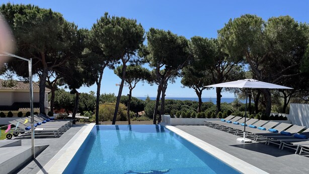 Une piscine rectangulaire à débordement, avec une eau d’un bleu cristallin, entourée de chaises longues et de parasols, le tout se fondant dans un décor de verdure et offrant une vue sur la mer.