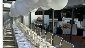 Une longue table à manger, recouverte de nappes blanches, agrémentée de verres et de compositions florales, sous une guirlande de ballons blancs.