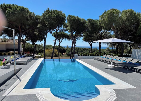 A rectangular infinity pool with clear blue water, surrounded by lounge chairs and umbrellas, set against a backdrop of trees and a view of the sea.
