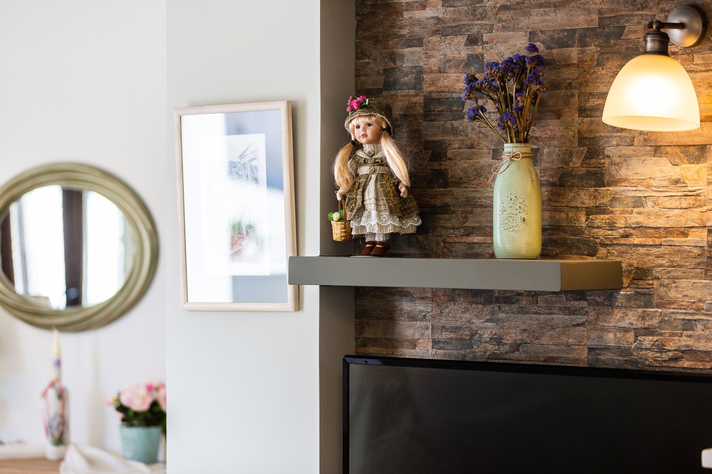 A doll on a shelf, a vase with flowers, and a wall-mounted lamp.