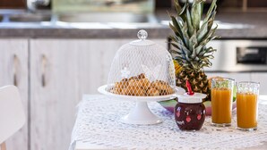 Disposição para o pequeno-almoço com uma abacaxi, uma tigela de cereais com tampa, um frasco de geleia e dois copos de sumo de laranja sobre uma mesa com toalha bordada.