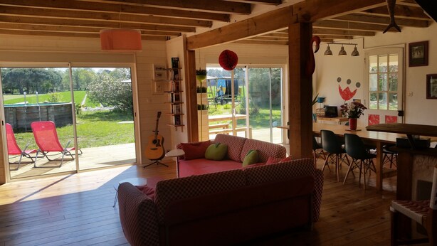 Ein geräumiges Wohnzimmer mit Holzboden, einem roten Couch mit Muster und einer Gitarre. Es gibt einen Essbereich mit Tisch und Stühlen, und durch eine Glastür hat man einen Blick auf einen Garten.