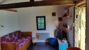 Ein gemütliches Zimmer mit Holzbalkendecke, einer Couch mit gemustertem Bezug, einem Fernseher, einem kleinen Regal mit Dekoration und einem Fenster mit Blick ins Grüne.