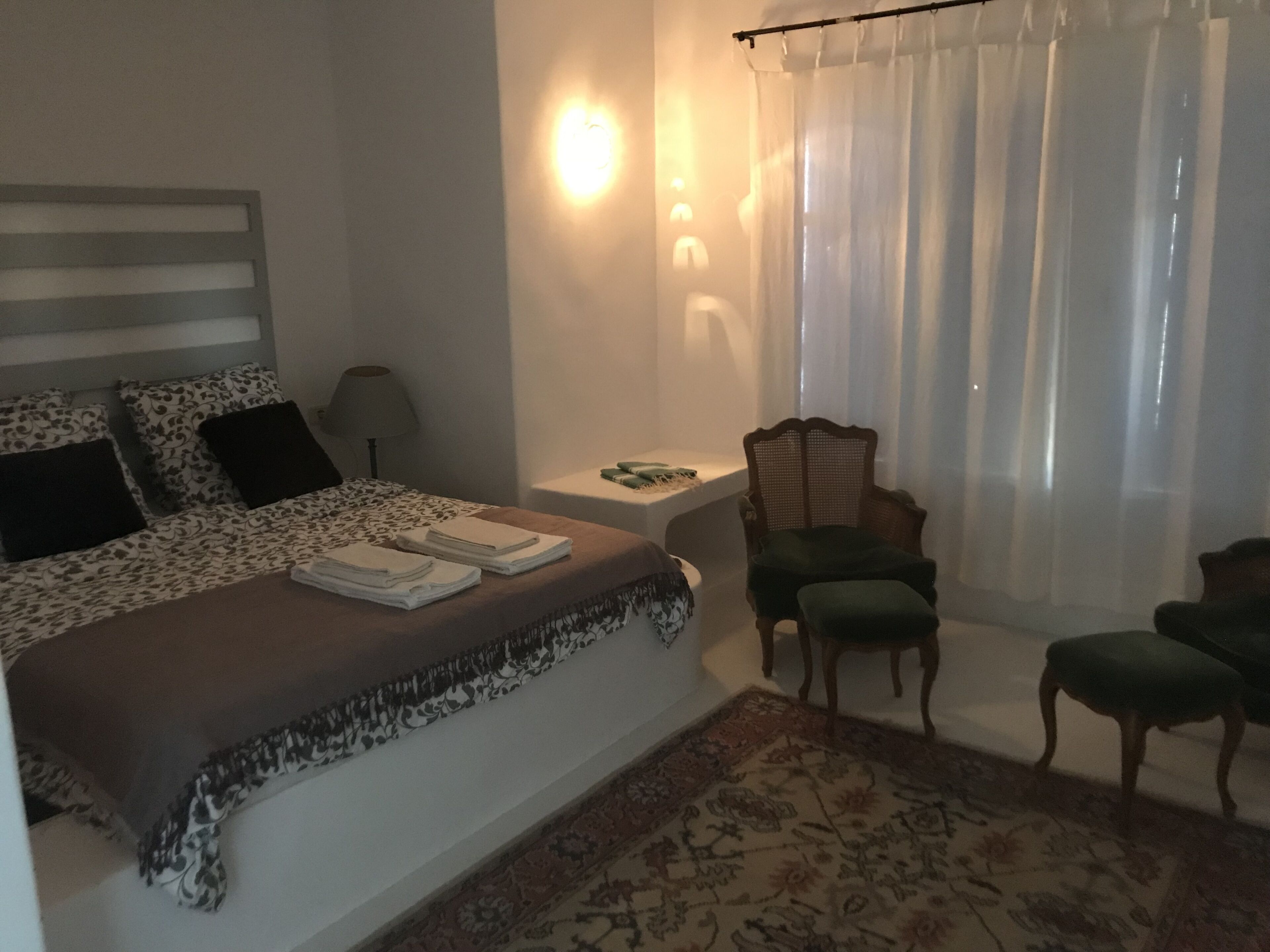 Et soveværelse med seng, stol, lille bord og et vindue med gardiner.