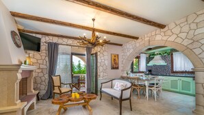 A spacious living area with a stone wall, wooden furniture, and a kitchen visible through an archway. - Fairytale Villa, in the heart of Lefkas town and Agios Ioannis beach! (Lefkada)