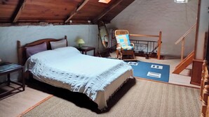 A cozy attic bedroom with a wooden ceiling, a bed with a white bedspread, a rocking chair, and a small table with a lamp. - LA GRANGE A CONVERTED BARN IN THE BEAUTIFUL HAMLET OF TALROCH (Melrand)