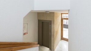 En smal hall med en trätrappa på vänster sida, en beige hiss i mitten och en trädörr längst ner.