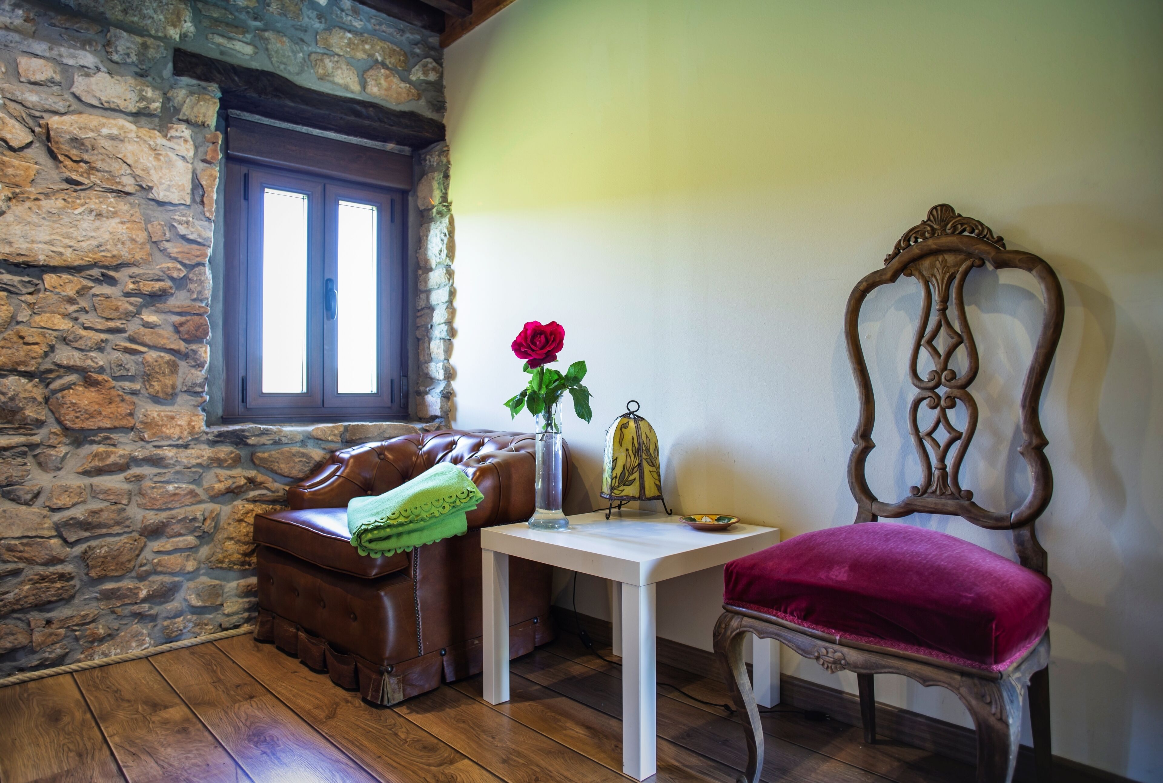 Una habitación con pared de piedra, un banco de madera, una mesa con un jarrón de flores y una silla con cojín.