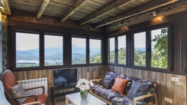 A cozy room with a wooden ceiling, large windows offering a view of mountains, a sofa with patterned cushions, a chair, and a TV on a stand. - Ruralmédulas Las Médulas - El Bierzo house 4 (Carucedo)