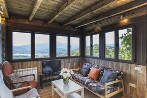 Ein gemütliches Zimmer mit Holzboden, großen Fenstern mit Bergblick, einem Sofa mit gemusterten Kissen, einem Sessel und einem Fernseher auf einem Schrank.