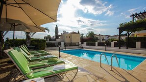 Ein Swimmingpool mit Liegestühlen und Sonnenschirmen, umgeben von einer Steinterrasse und Blick auf Gebäude im Hintergrund.