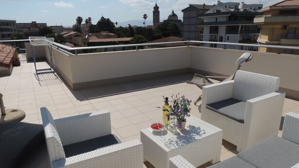 A rooftop terrace with white wicker furniture, a satellite dish, and a view of buildings and a tower in the background. - Top floor penthouse apartment with terrace in the historical center, 3 km from the beach (Oristano)