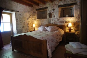 Traditional stone walls and rustic wooden furniture create a cozy atmosphere, featuring a plush bed with a floral-patterned bedspread and two pillows.