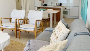 Ein modernes Wohnzimmer mit einem grauen Sofa, Holzstühlen, einem kleinen runden Tisch und einer Küche im Hintergrund.