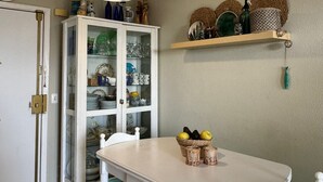 Et spiseområde med et hvidt bord og stole, et skab med glaslåger samt en vægmonteret hylde med pynt.