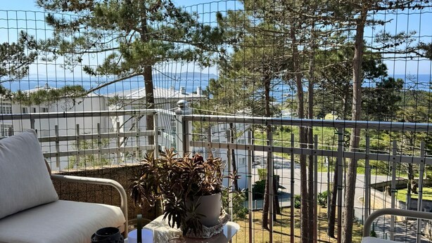 En balkon med en sofa, et lille bord med en plante, og udsigt til havet og træerne.