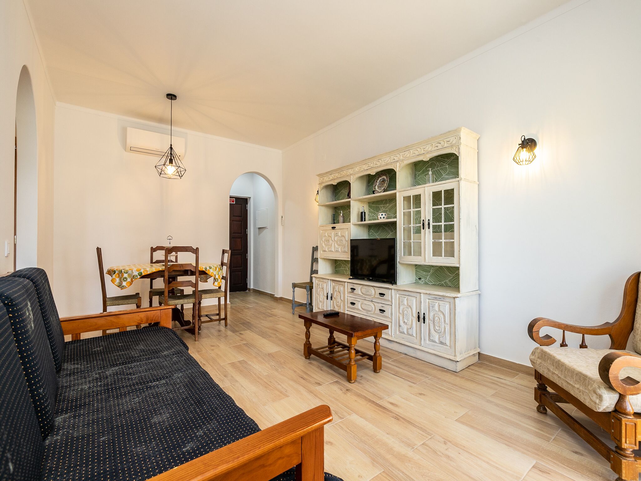 Un salon avec un canapé, une table à manger avec des chaises, un meuble TV avec des placards et un couloir avec une arche.