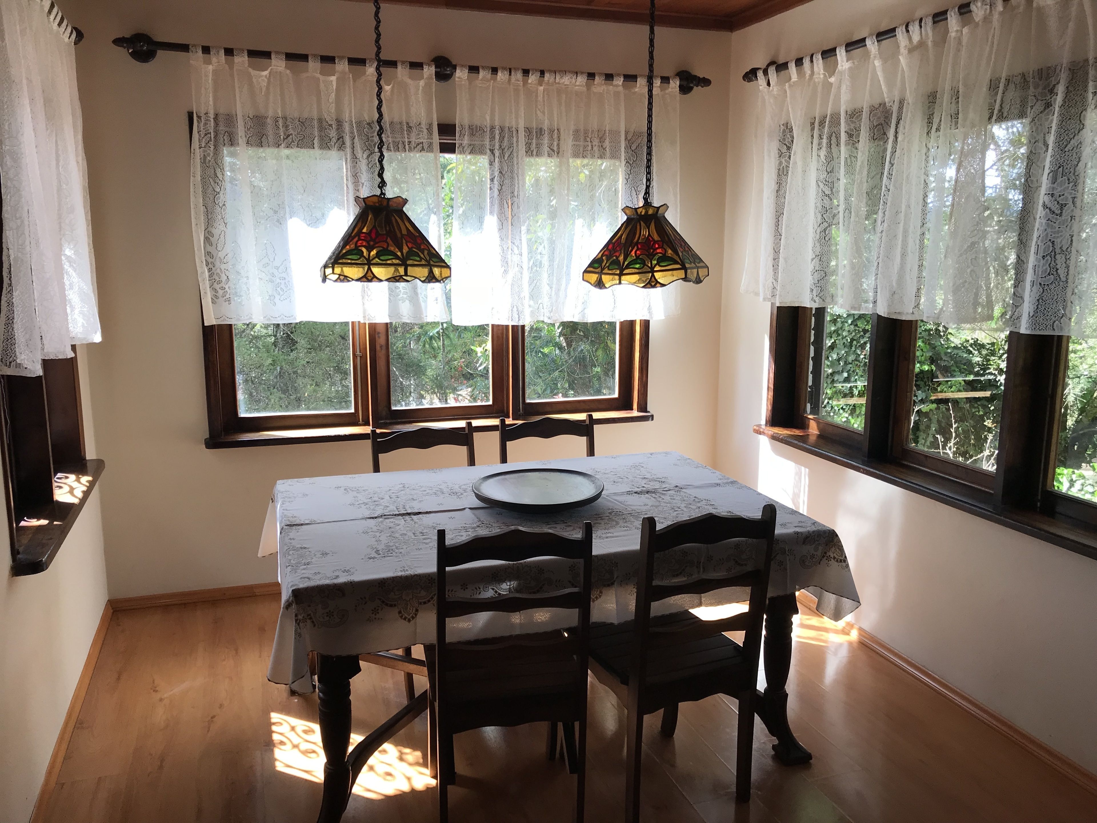 A dining area with a table set for two, two hanging lamps, lace curtains, and windows with a view of greenery.