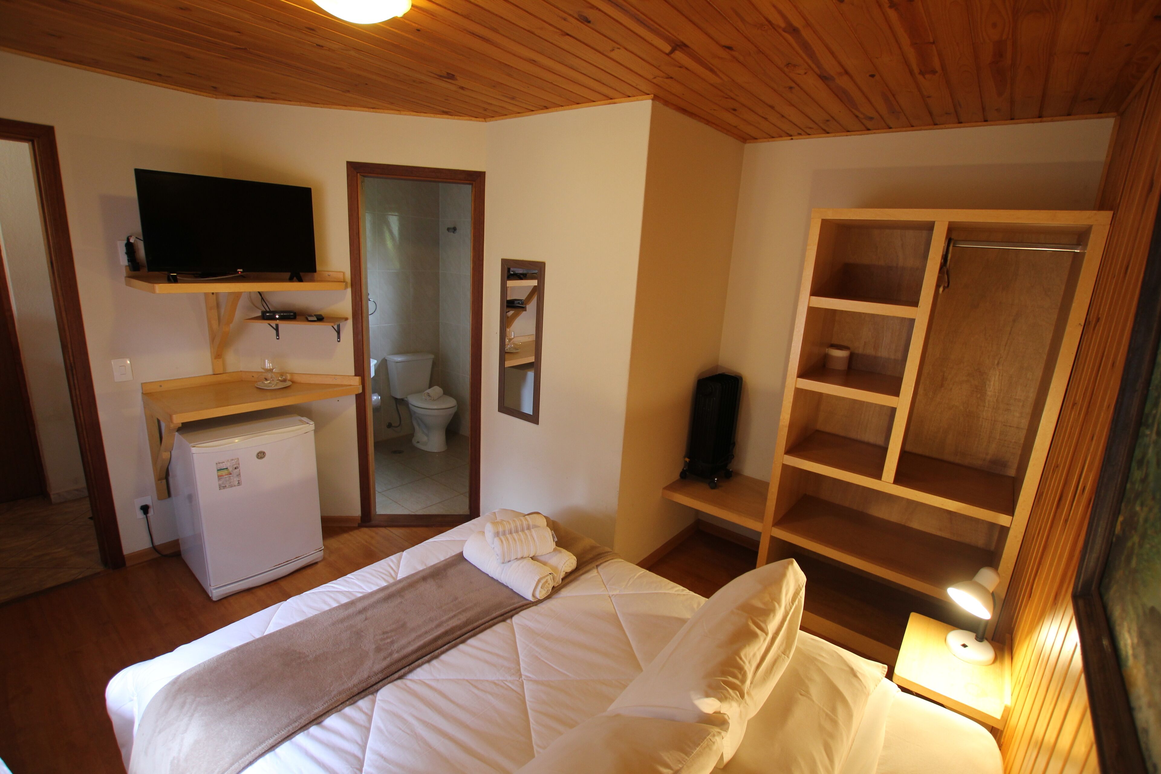 Un dormitorio con cama, televisor, refrigerador pequeño, espejo y armario de madera.