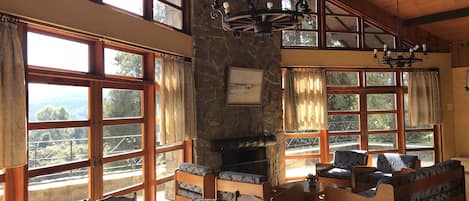 A cozy room with a stone fireplace, wooden furniture, and large windows offering a view of the outdoors.
