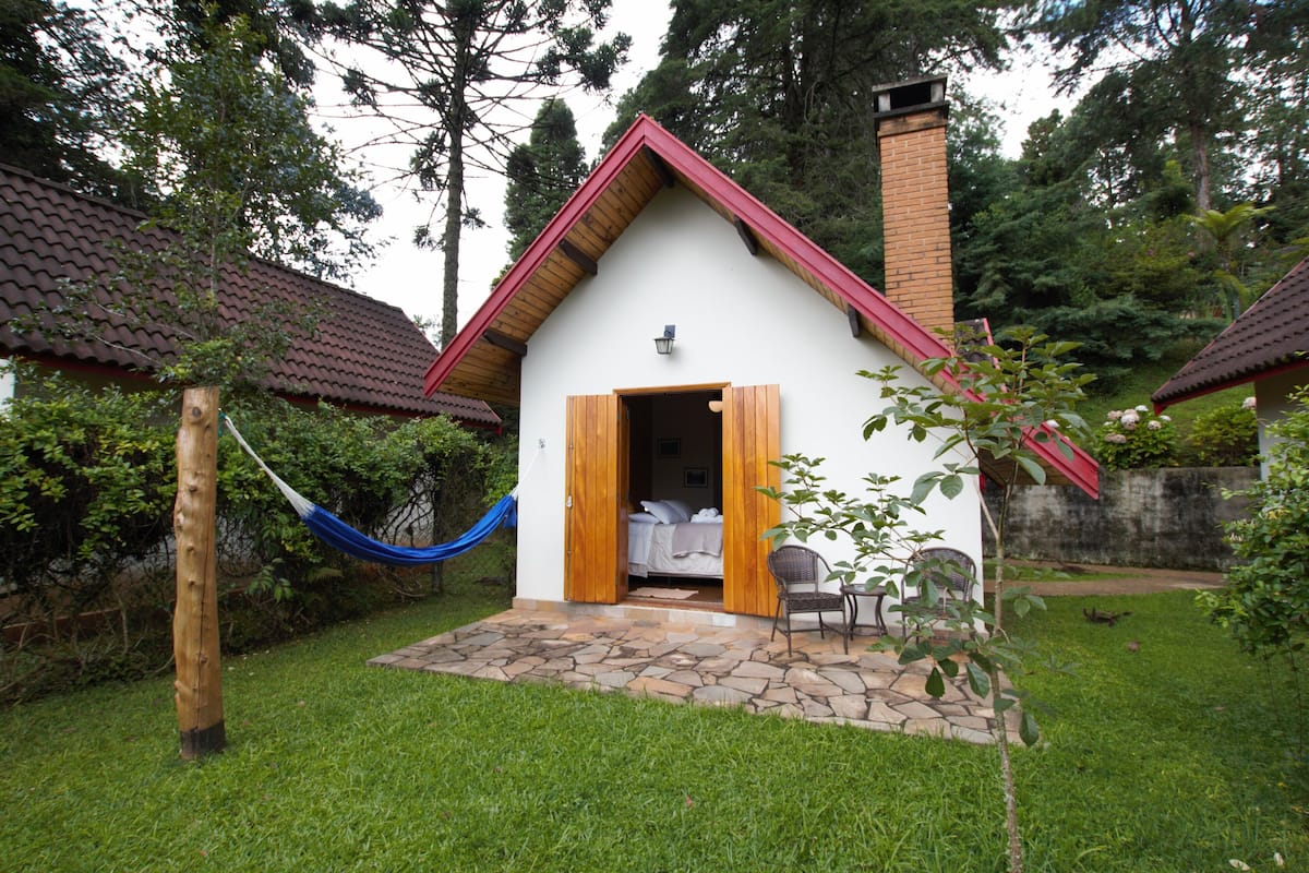 Ein kleines, eingeschossiges Haus mit rotem Dach und Schornstein, einer Hängematte und einer Terrasse mit Tisch und Stühlen.