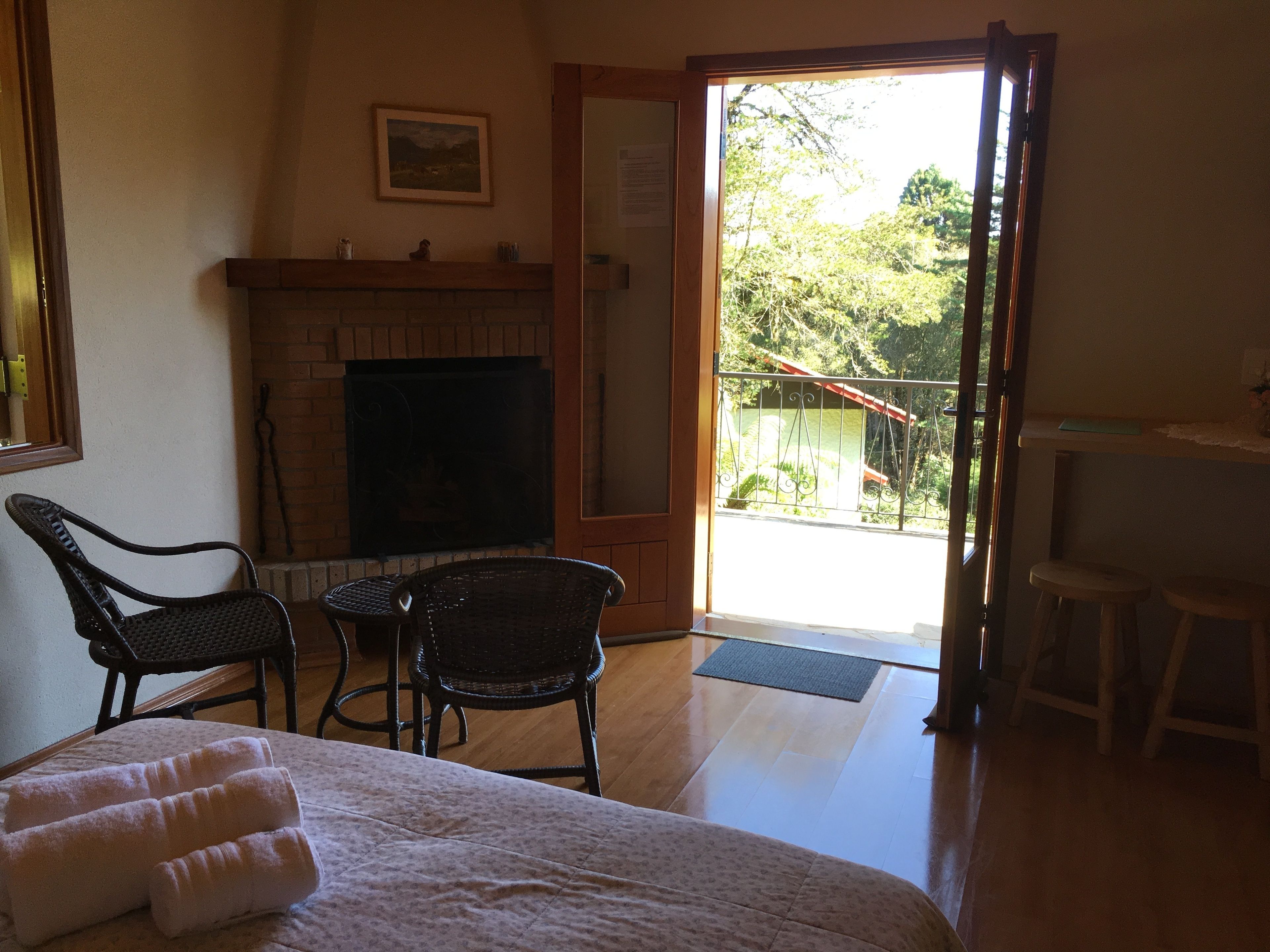 A room with a fireplace, wicker chairs, a bed with towels, and a balcony with a view of greenery.