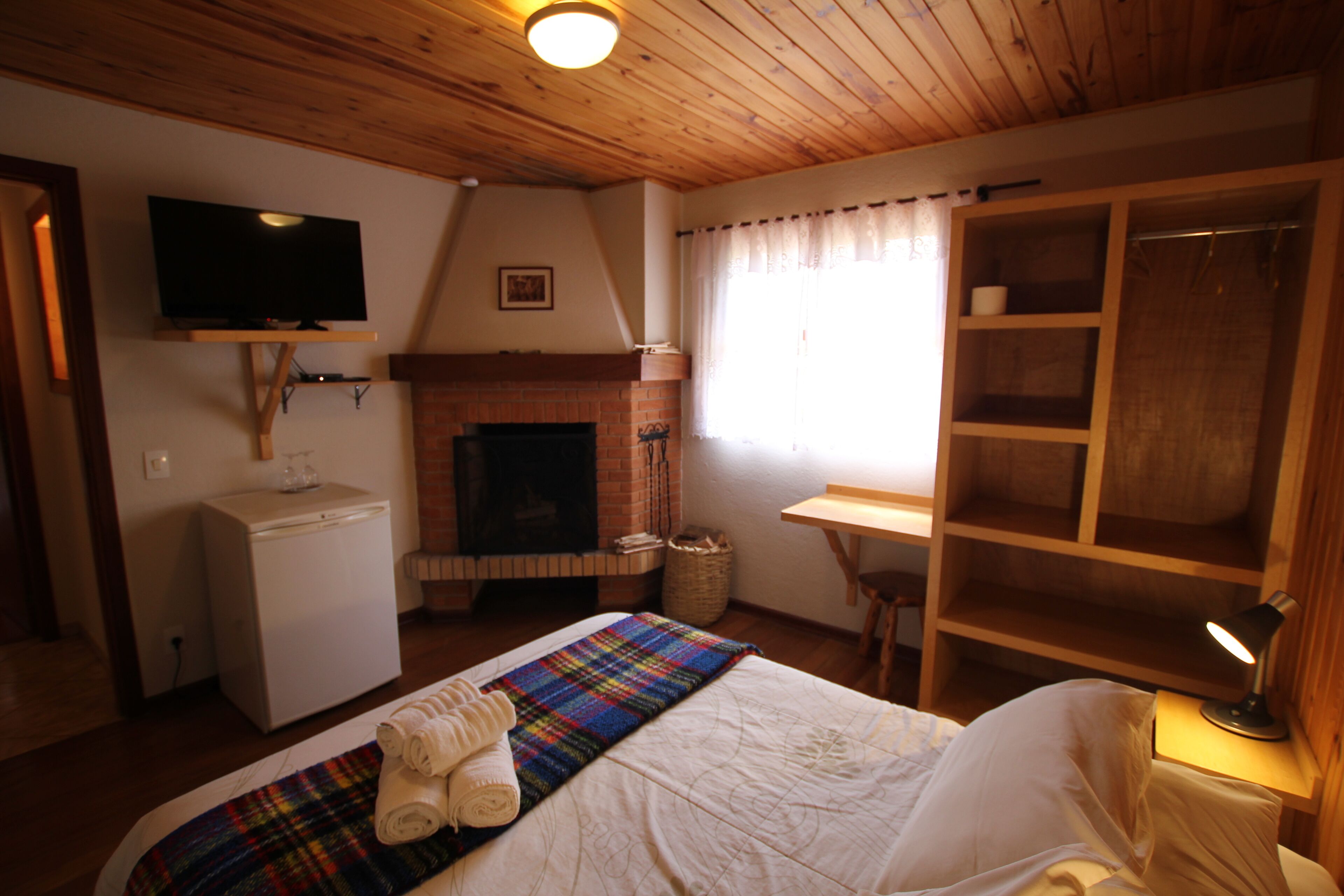 A cozy room with a bed, a television, a fireplace, a wooden wardrobe, and a desk with a lamp.