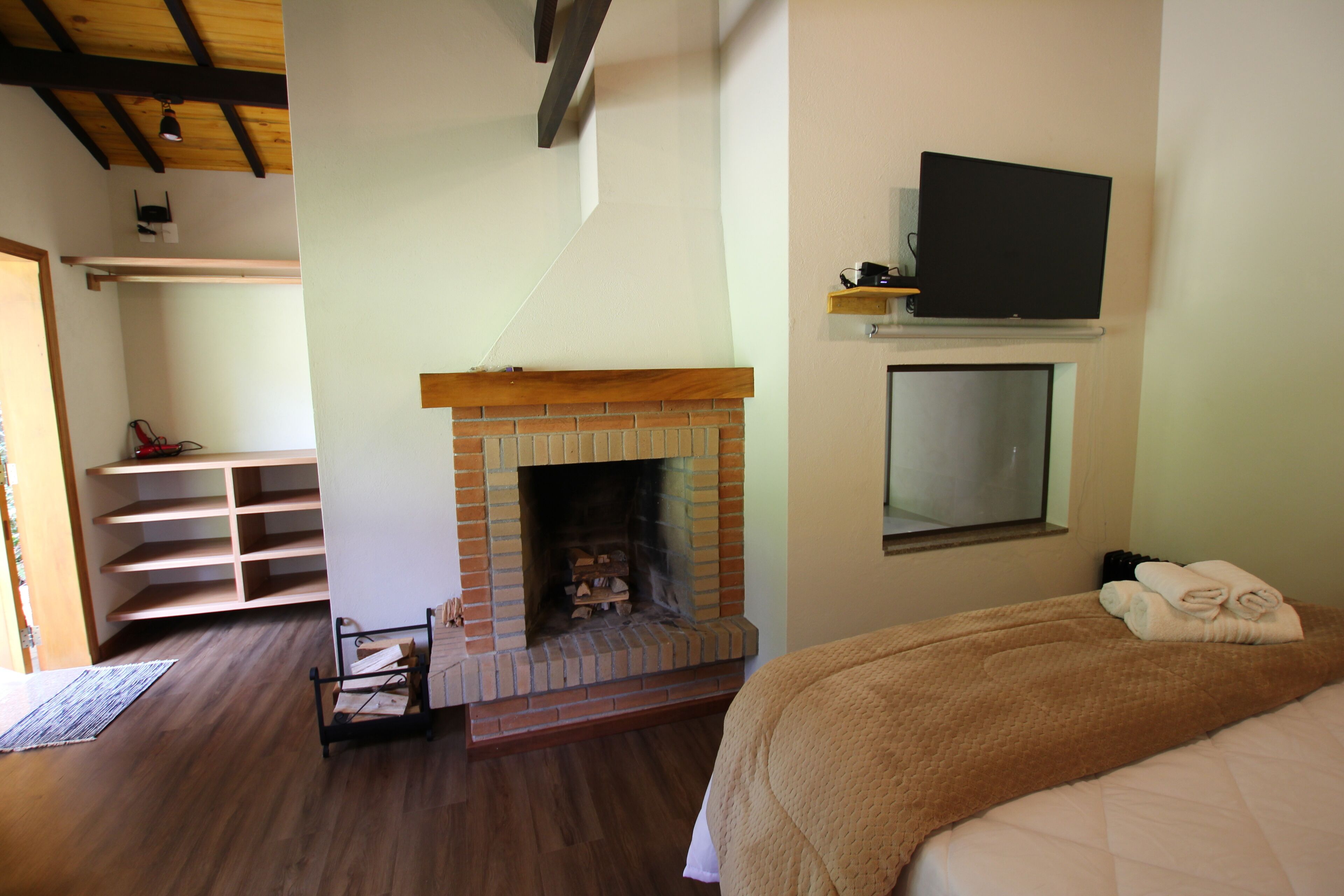 A bedroom with a brick fireplace, a bed with a brown blanket, a TV mounted above the fireplace, and a wooden ceiling.