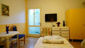 Quarto com cama, televisão, cômoda, cadeira, mesa e porta que dá para uma varanda.