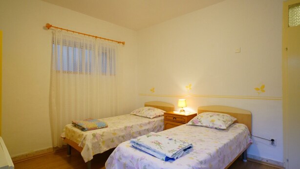 Quarto com duas camas de solteiro, uma mesinha de cabeceira com abajur, uma janela com cortinas e uma porta.