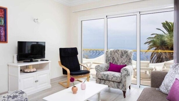 A living room with a television, a sofa, a chair, and a table. There is a balcony with a view of the sea. - Cozy home in Tenerife with views & pool (Santa Cruz de Tenerife)