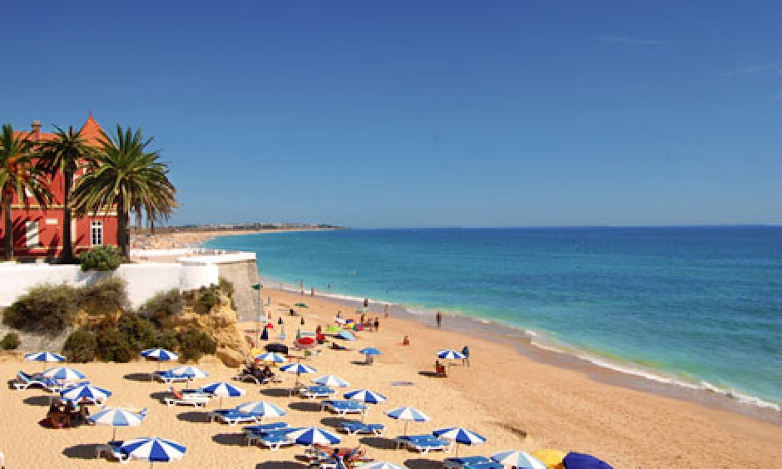 A beach with numerous blue and white umbrellas, a sandy shore, and a red building with a red roof.