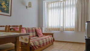 Um quarto aconchegante com uma cama de madeira, uma mesinha, uma luminária na parede e uma janela com cortinas leves.