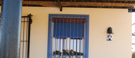 A covered outdoor seating area with a bench and chairs, a window with blue shutters, and a wall-mounted light fixture.
