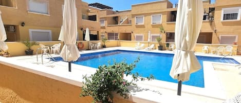 A swimming pool with white umbrellas, a small plant, and a building with multiple balconies.