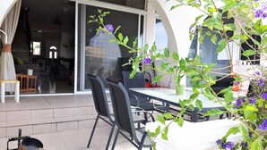An outdoor patio with a table and chairs, a sliding glass door leading to an interior space, and a plant with purple flowers.