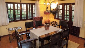 Une salle à manger avec une table en bois, des chaises et un lustre. On y trouve des fenêtres avec des rideaux, un vase rempli de fleurs et un jeu d’échecs posé sur la table.
