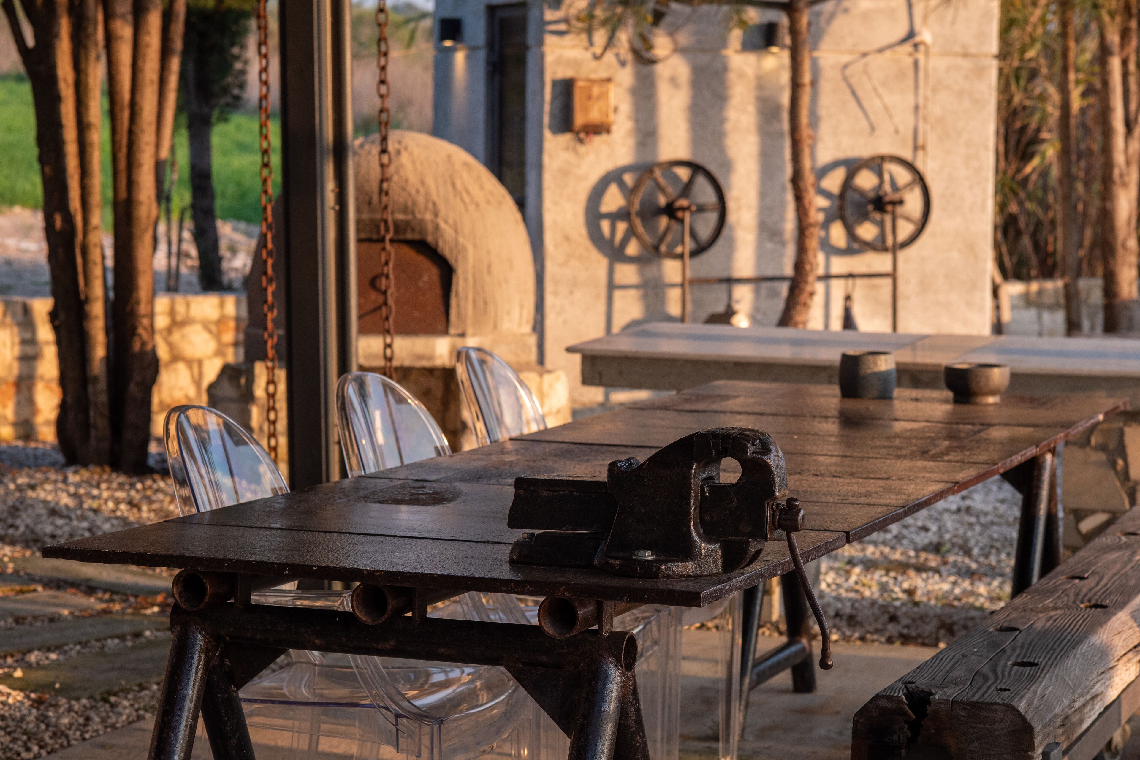 A rustic outdoor dining area with a wooden table, metal chairs, and a stone oven in the background.