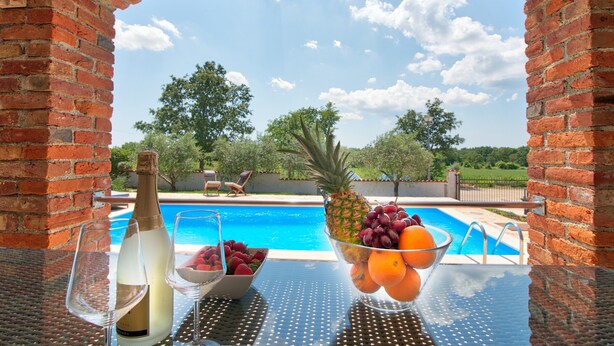 Ein Tisch am Pool mit einer Champagnerflasche, einer Obstschale und einem Weinglas, mit Blick auf ein Schwimmbecken unter blauem Himmel.